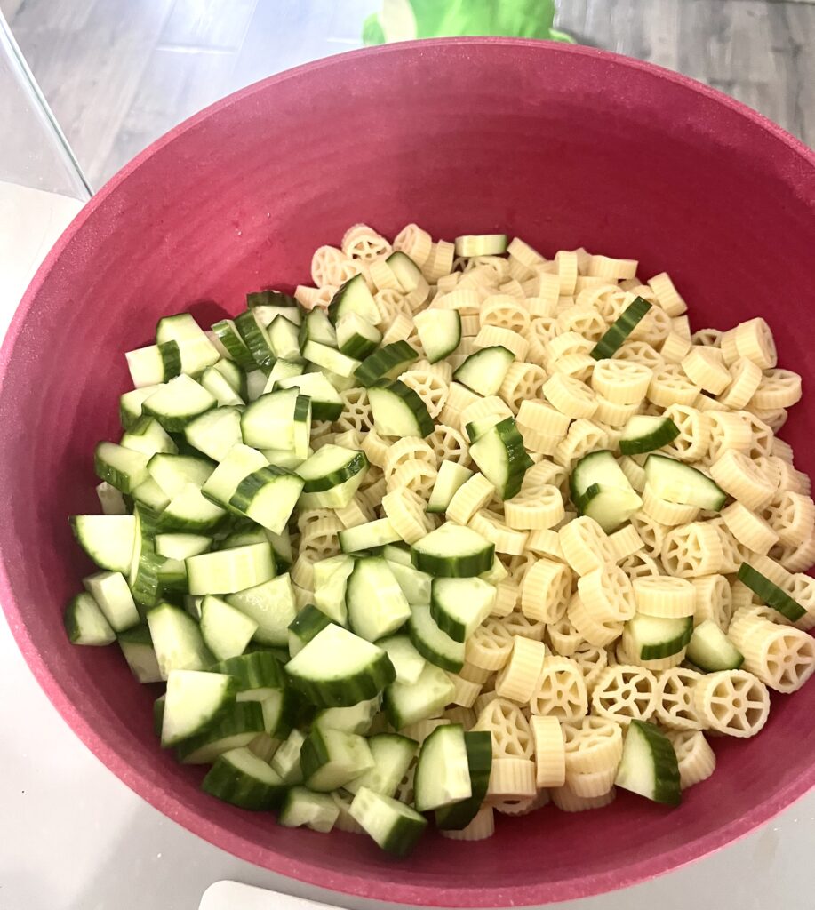 Cooked wheel pasta and chopped cucumbers in a red bowl, ready to be mixed for an easy pasta salad recipe