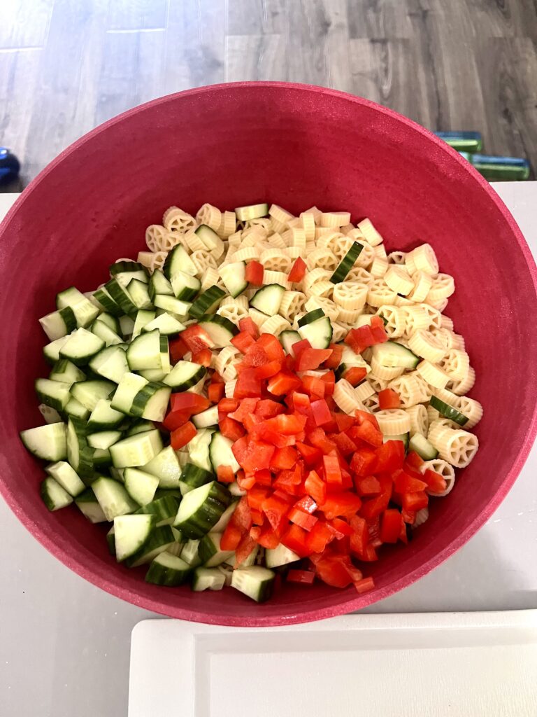 Wheel pasta, chopped cucumbers, and diced red bell peppers layered in a red bowl for a fresh and easy pasta salad recipe