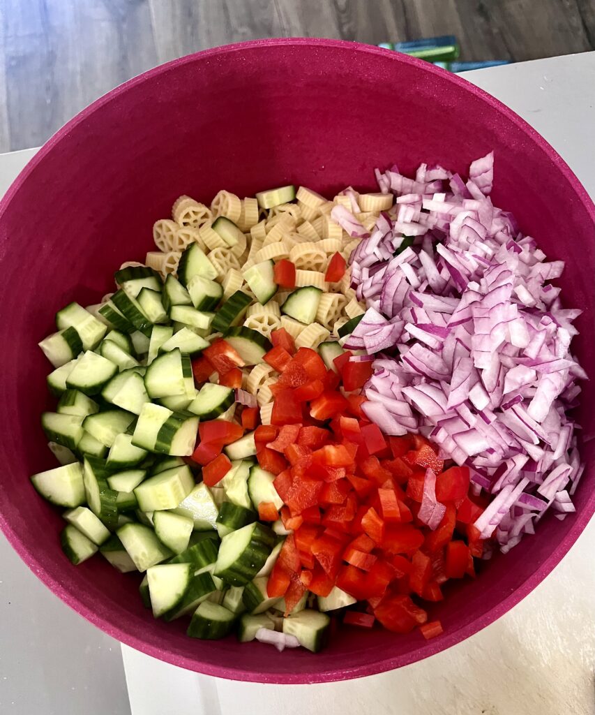 Unmixed pasta salad ingredients in a red bowl, including wheel pasta, cucumbers, red bell peppers, and chopped red onion