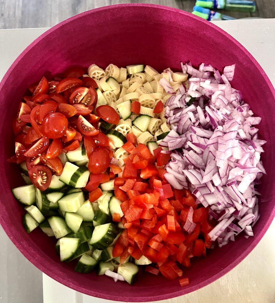 Colorful pasta salad ingredients in a red bowl, including cherry tomatoes, cucumbers, red bell peppers, red onion, and wheel pasta