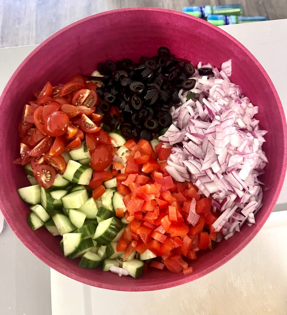 Unmixed easy pasta salad ingredients in a red bowl, including cherry tomatoes, cucumbers, red bell peppers, red onion, and sliced black olives
