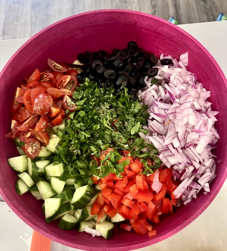 Layered pasta salad ingredients in a red bowl, including cherry tomatoes, cucumbers, red bell peppers, red onion, black olives, and fresh chopped parsley