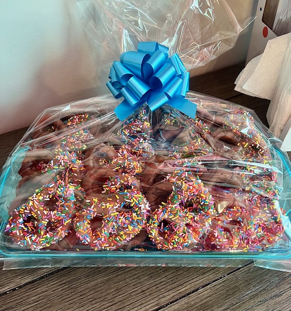 Tray of chocolate-covered pretzels topped with rainbow sprinkles, wrapped in clear cellophane and tied with a blue bow, served as a festive treat at a carnival birthday party