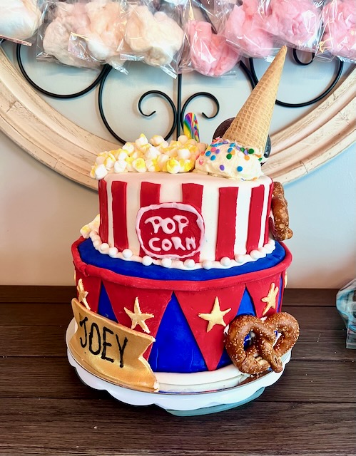 Two-tiered carnival birthday cake decorated with circus tent designs, popcorn, ice cream cone, soft pretzel, and a personalized name banner, perfect for a carnival party dessert table.