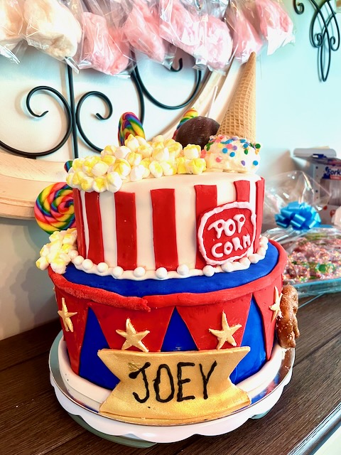 Side view of a vibrant carnival-themed birthday cake with popcorn topping, candy decorations, an ice cream cone, and custom name banner, ideal for a circus or carnival party dessert display.