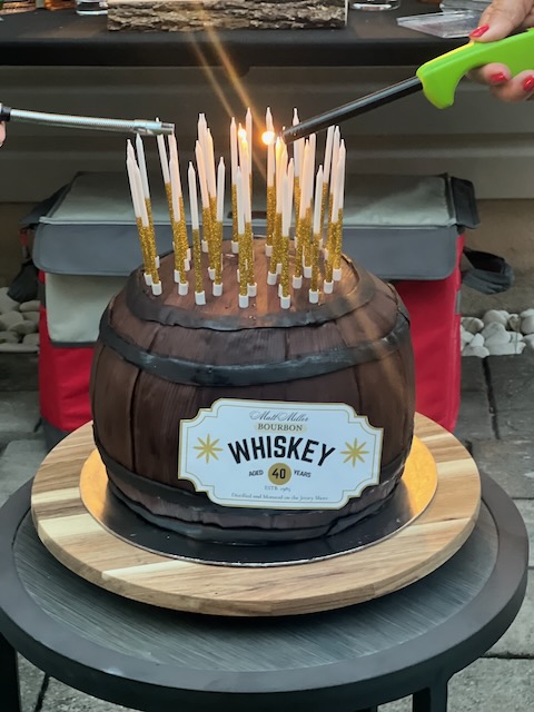 Whiskey barrel-shaped birthday cake decorated with tall white and gold candles being lit for a Bourbon and BBQ themed 40th birthday celebration.
