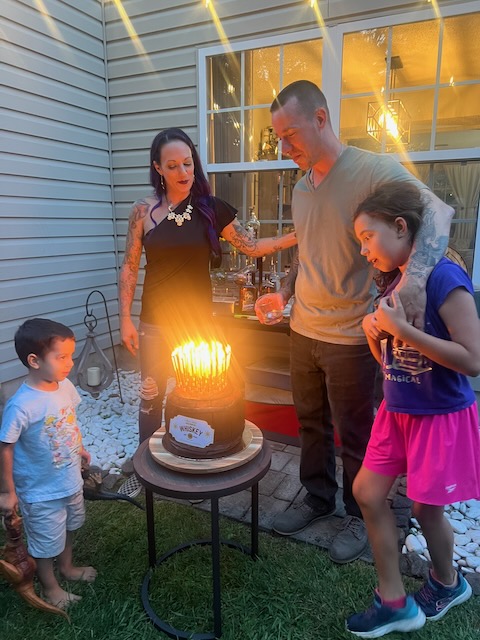 Birthday boy celebrating his 40th with family gathered around a whiskey barrel cake glowing with tall golden candles at a Bourbon and BBQ party