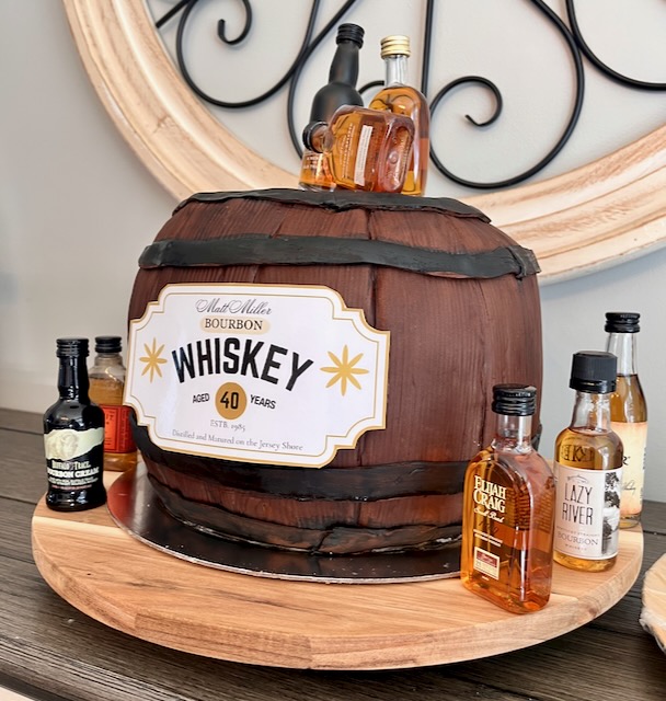 Whiskey barrel birthday cake decorated for a Bourbon & BBQ 40th birthday party, with maple bourbon flavors, custom label, and mini bourbon bottles on top.