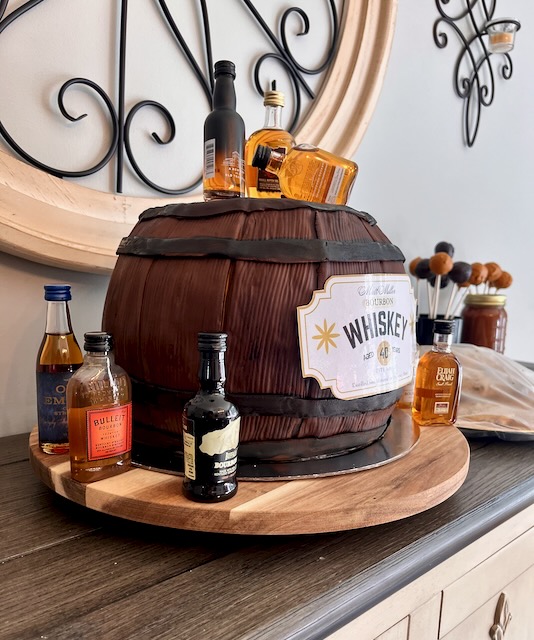 Side view of whiskey barrel birthday cake for a Bourbon & BBQ 40th birthday party, decorated with a custom label and surrounded by mini bourbon bottles.