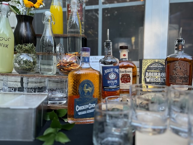 Self-serve bourbon bar setup at a Bourbon & BBQ 40th birthday party with assorted bourbon bottles, custom rocks glasses, sour mix, simple syrup, and garnishes.
