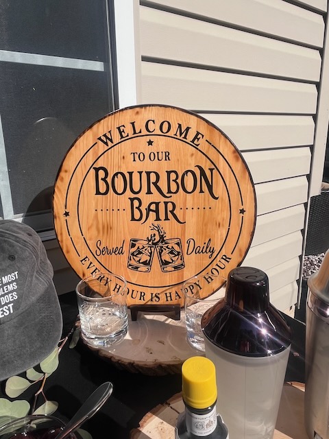 Wooden Bourbon Bar sign displayed at a 40th birthday party, engraved with ‘Welcome to our Bourbon Bar, Served Daily, Every Hour is Happy Hour,’ styled with cocktail shakers and glasses.