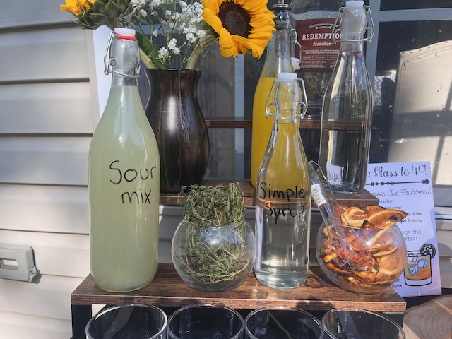 Bourbon bar detail with bottles of homemade sour mix and simple syrup, fresh herbs, dried orange slices, and sunflowers at a Bourbon & BBQ 40th birthday party.