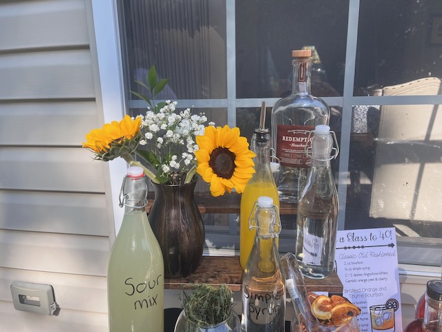 Close-up of bourbon bar setup at a Bourbon & BBQ 40th birthday party with sunflowers in a vase, sour mix, simple syrup, bourbon bottles, and a signature cocktail sign.
