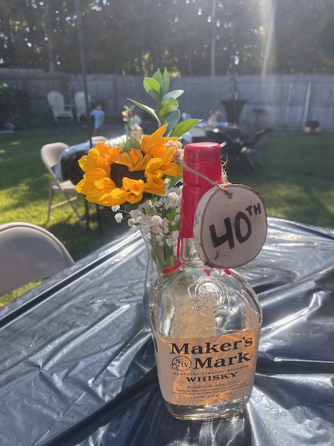 DIY centerpiece made from an empty Maker’s Mark bourbon bottle with sunflowers and greenery, decorated with a wooden 40th birthday tag at a Bourbon & BBQ party.