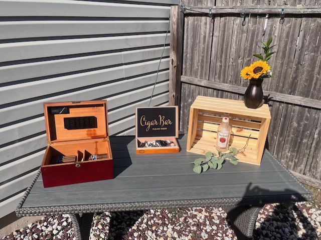 Outdoor cigar bar setup at a 40th birthday Bourbon and BBQ party, featuring a humidor, cigar sign, and rustic decor with flowers in a bourbon bottle vase.