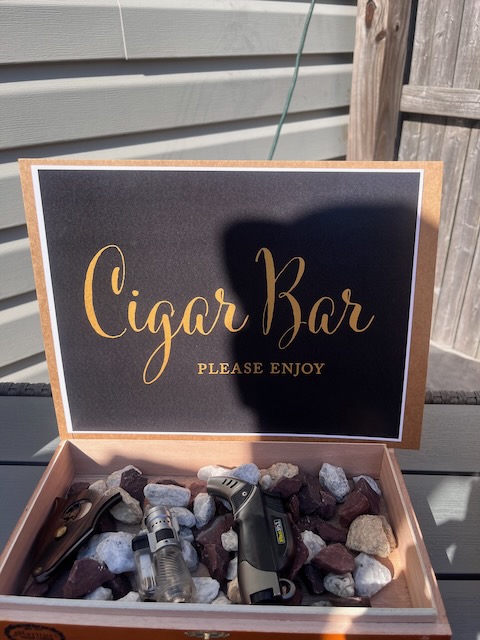 Close-up of a cigar bar sign with gold lettering reading ‘Cigar Bar Please Enjoy,’ displayed with lighters and cutters in a rustic box at a 40th birthday Bourbon and BBQ party.