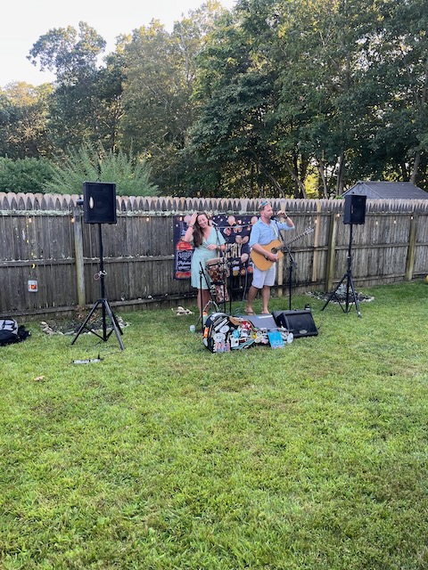 Live acoustic band performing in the backyard at a 40th birthday Bourbon and BBQ party, with guitar, microphone, and speakers set up on the grass.