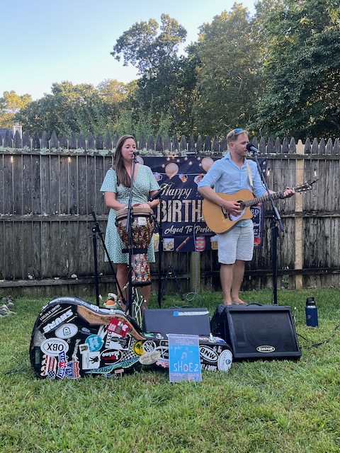 Acoustic duo performing live music with guitar and hand drum at a backyard 40th birthday Bourbon and BBQ party, with a festive birthday banner in the background.