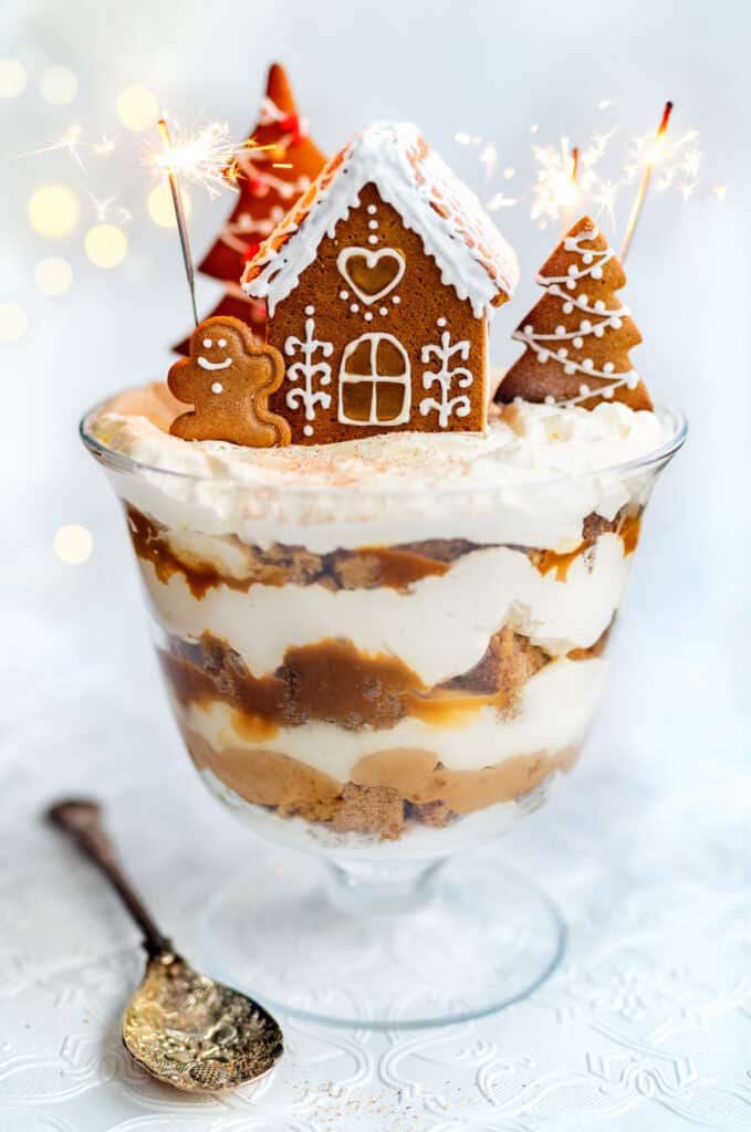 Gingerbread trifle layered with spiced cake, whipped cream, and caramel, topped with a gingerbread house and cookies in a glass dessert dish.