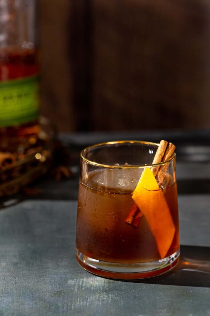Gingerbread old fashioned cocktail with cinnamon stick and orange peel garnish in a glass with a gold rim.